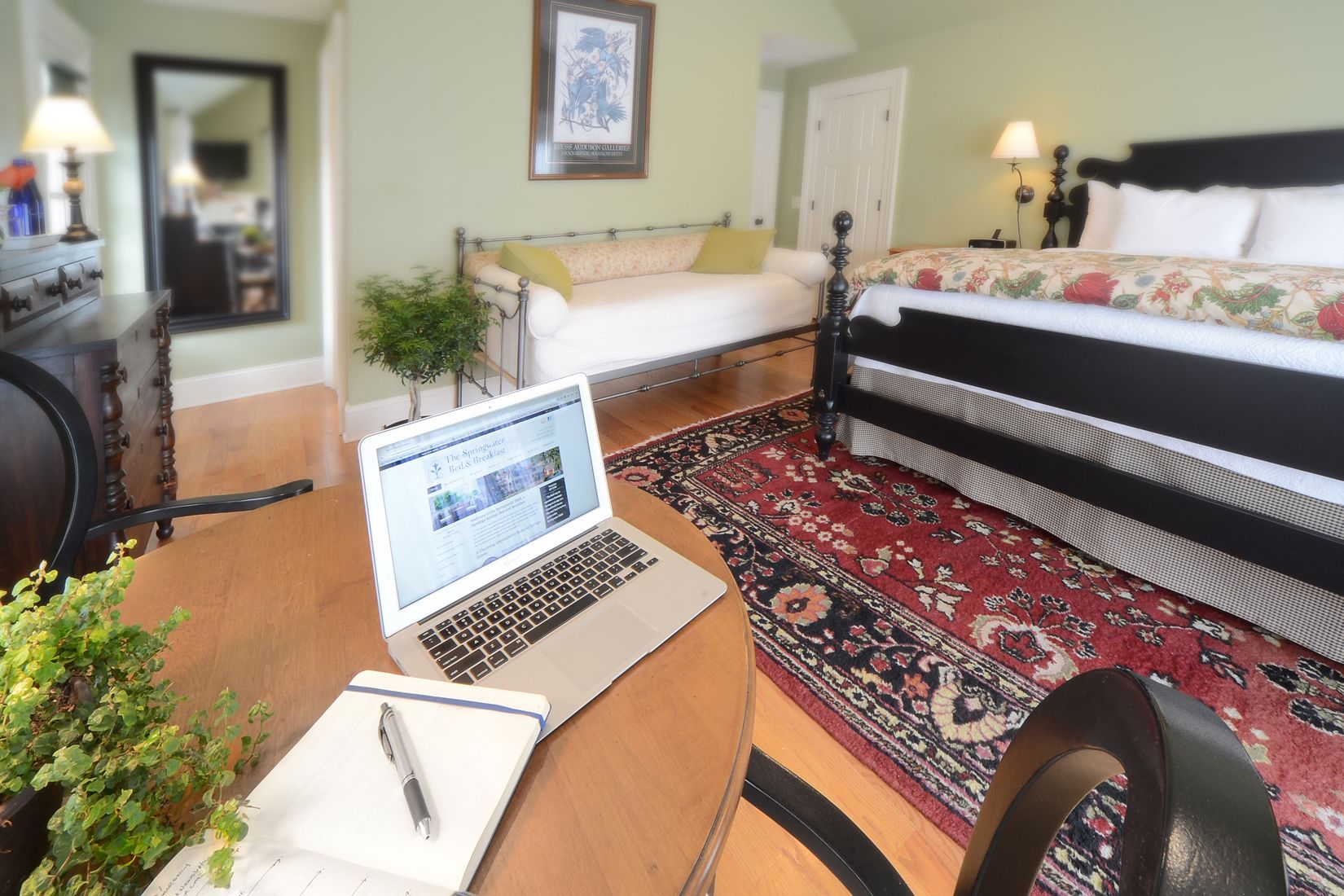 A hotel room with a bed, a desk with a laptop, a chair, a rug, and a plant.