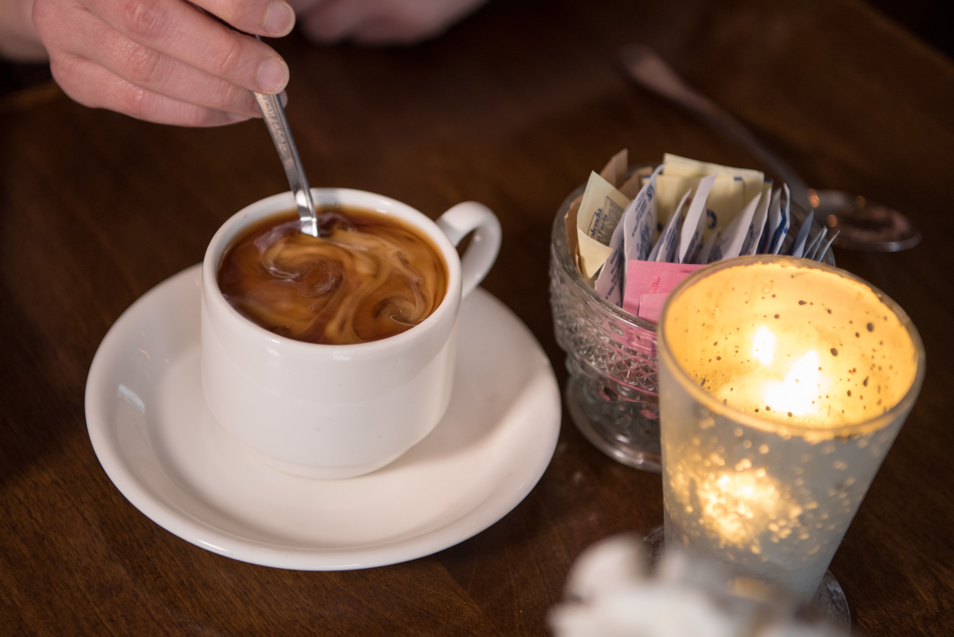 A cup of coffee on a saucer with a spoon, a glass candle holder, and a glass with a candle.