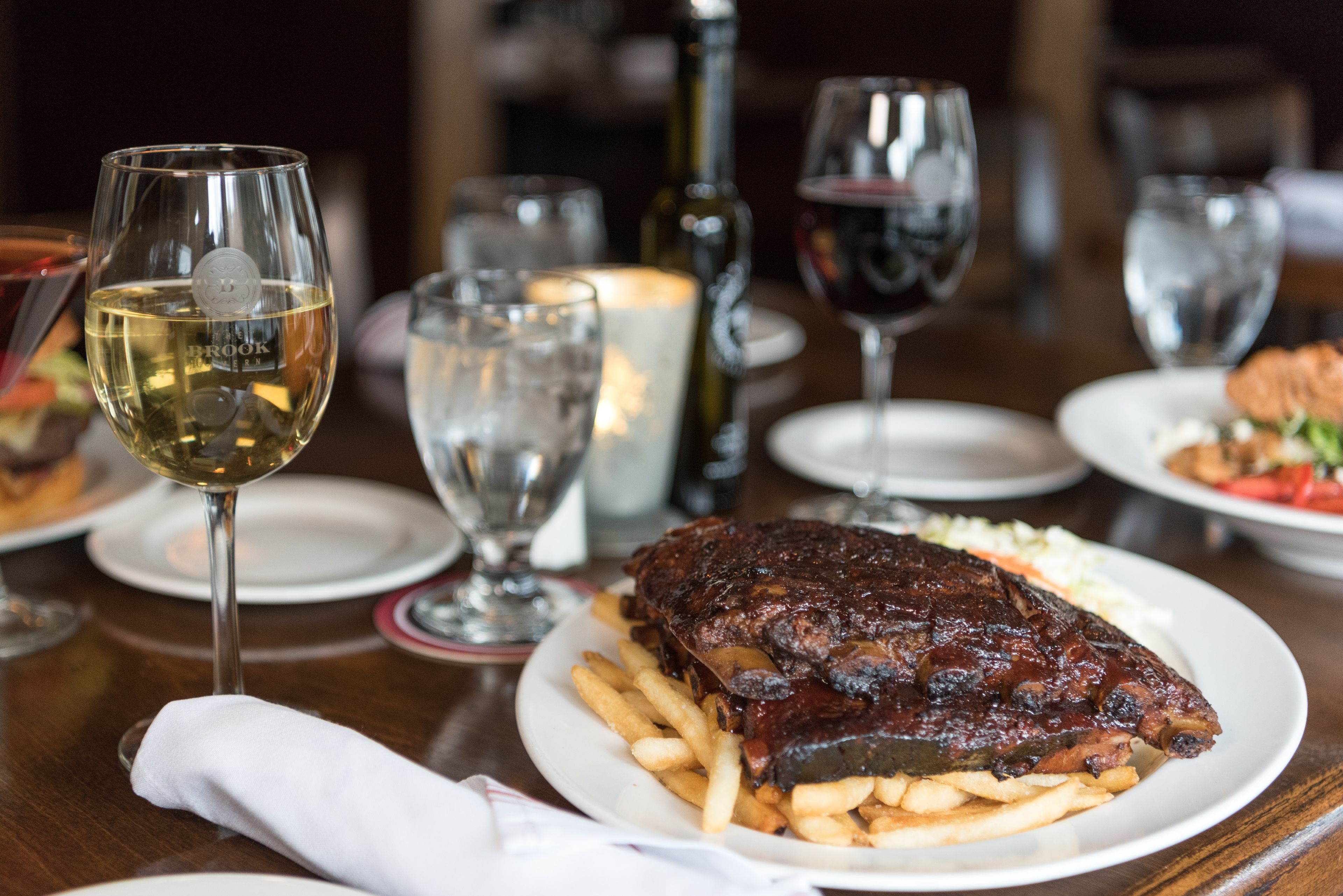 A dining table with a plate of ribs and fries, a bottle of wine, and several glasses.
