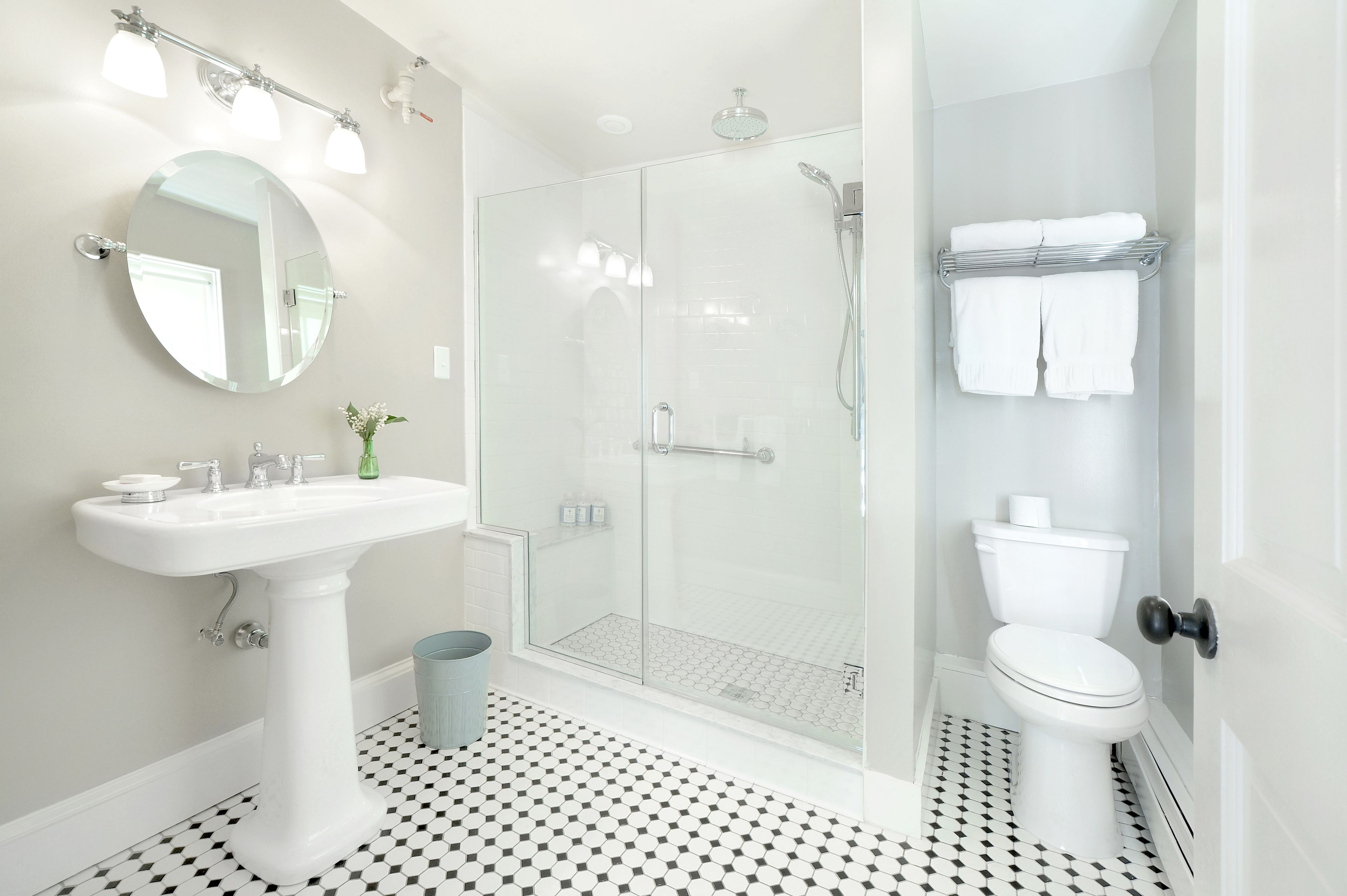 A bathroom with a pedestal sink, a round mirror, a glass-enclosed shower, a toilet, and a towel rack.