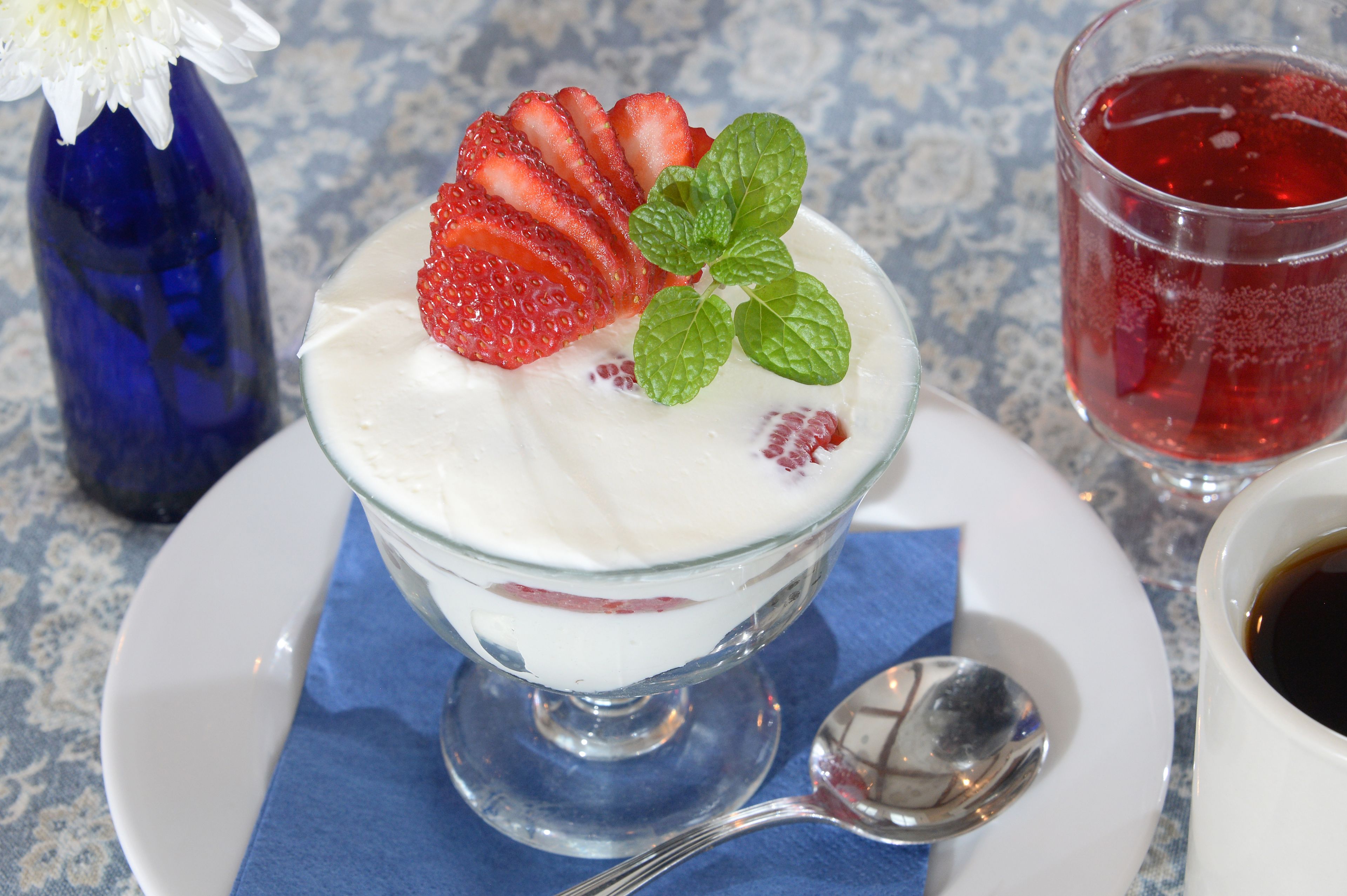A dessert in a glass bowl with strawberries and mint leaves on a white plate, accompanied by a spoon and a cup of coffee.