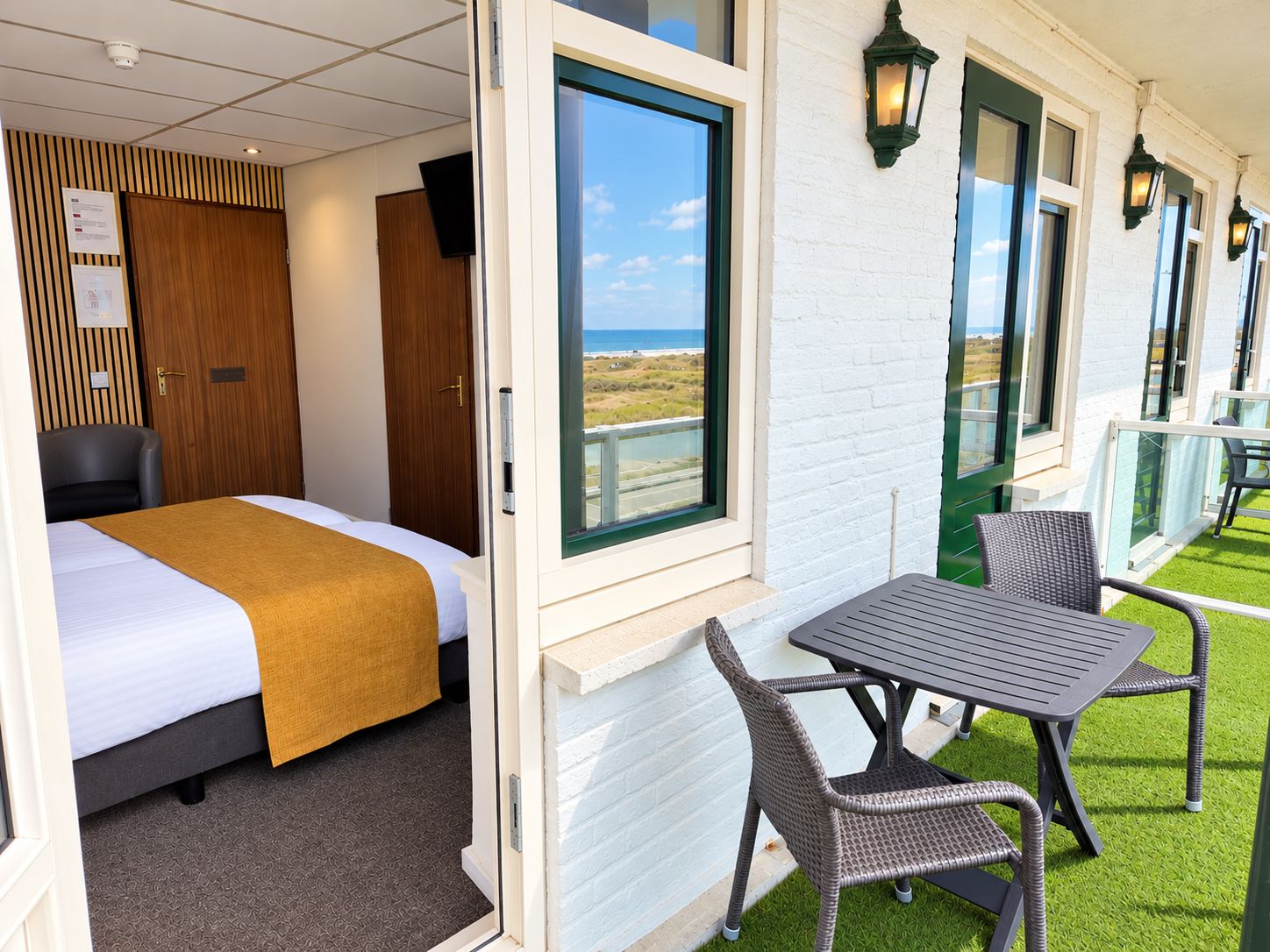 Een hotelkamer met een bed, een raam met uitzicht op het strand, een balkon met zitplaatsen buiten en een aan de muur bevestigde televisie.