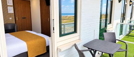 A hotel room with a bed, a window with a view of the beach, a balcony with outdoor seating, and a wall-mounted TV.