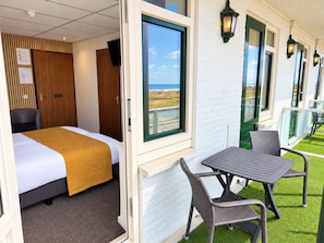 A hotel room with a bed, a window with a view of the beach, a balcony with outdoor seating, and a wall-mounted TV. - Hotel Zee en Duin (Katwijk Aan Zee)