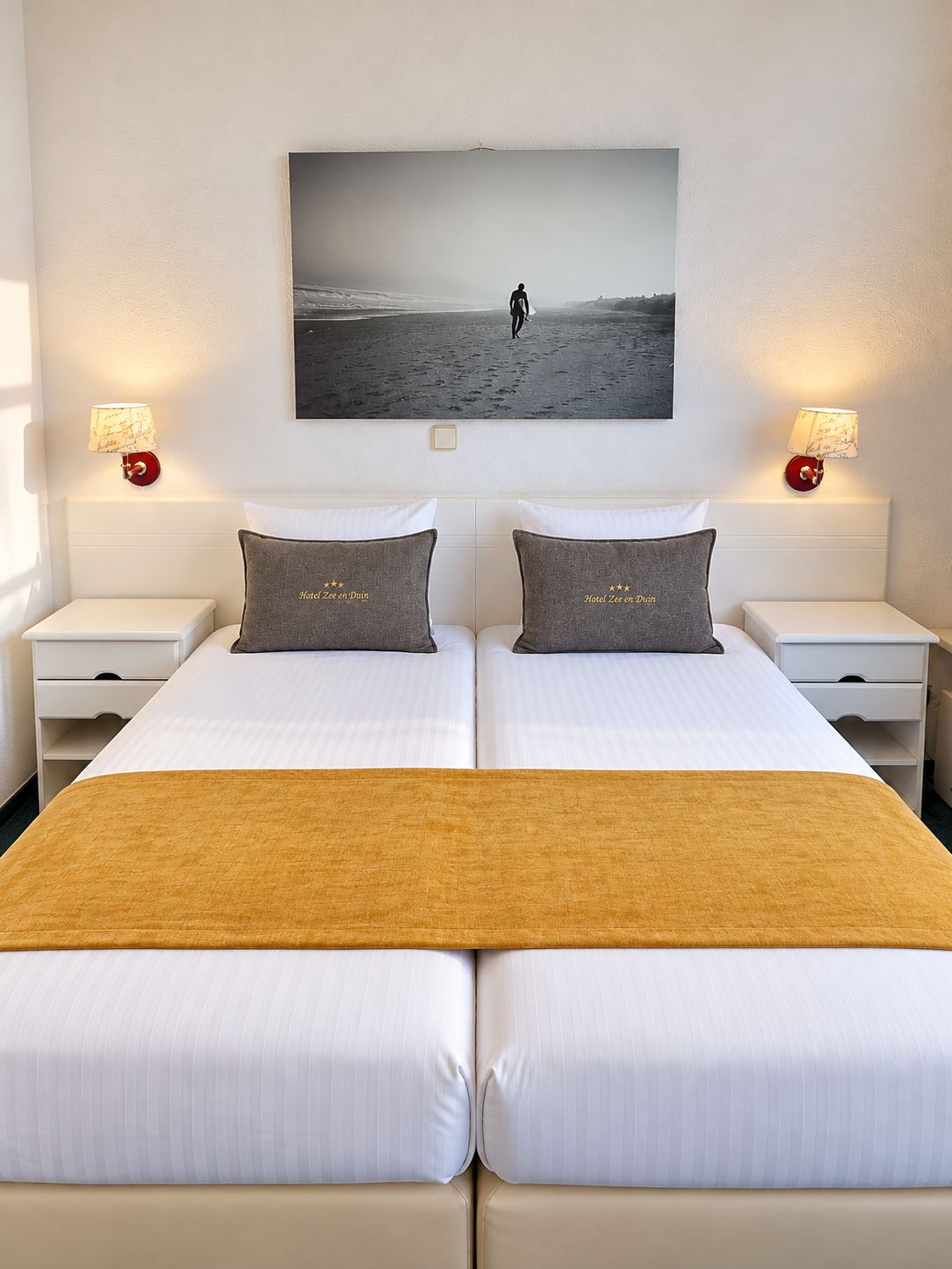 A double bed with a view of a beach scene, two bedside tables with lamps, and a single bed runner.