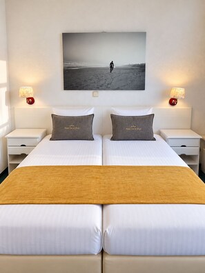 A double bed with a view of a beach scene, two bedside tables with lamps, and a single bed runner. - Hotel Zee en Duin (Katwijk Aan Zee)
