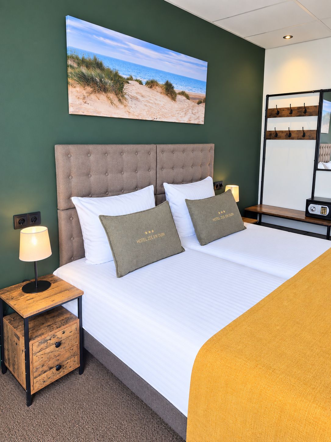 Habitación de hotel con cama, mesita de noche, lámpara, cajón, espejo fijado a la pared y un cuadro de playa.