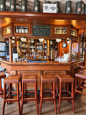 A bar area with wooden stools, a bar counter, and shelves stocked with bottles. - Hotel Zee en Duin (Katwijk Aan Zee)