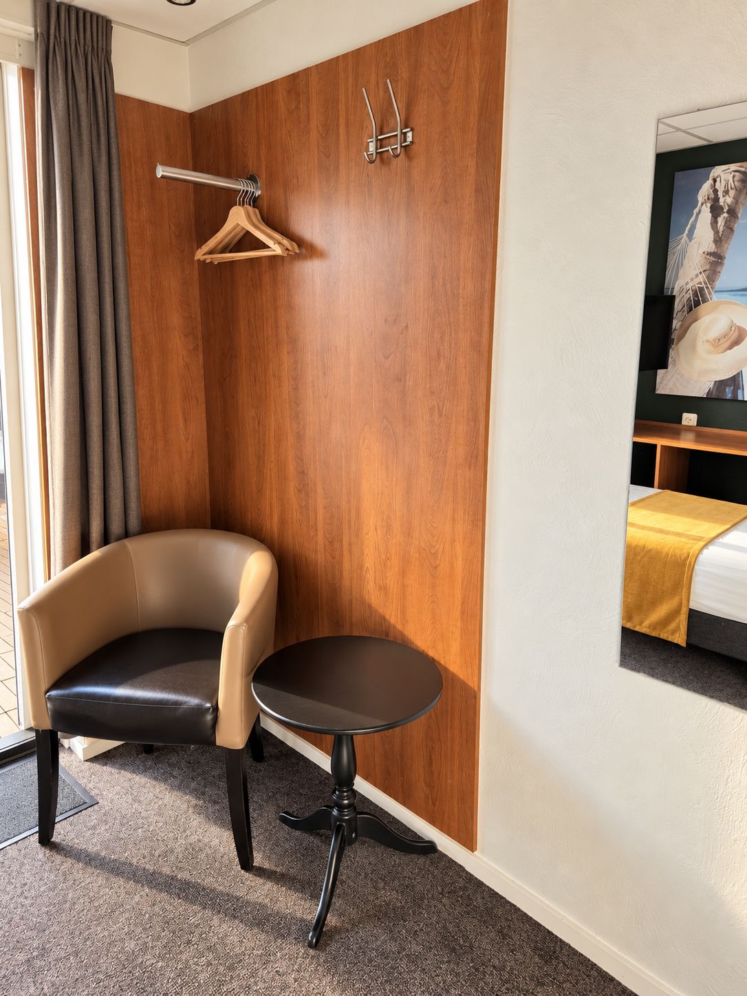 Een hotelkamer met een wand van houten panelen, een zwarte leren fauteuil, een klein rond tafeltje, een bed met een gele plaid en een wandtelevisie.