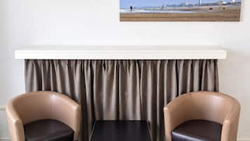 A room with a flat-screen TV mounted on the wall, a painting of a beach scene, a small table, two leather chairs, and a curtain.