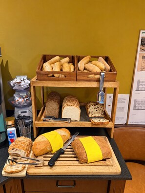 A variety of breads and pastries on a wooden display stand, a cutting board with bread and a knife, and a coffee machine in the background. - Hotel Zee en Duin (Katwijk Aan Zee)