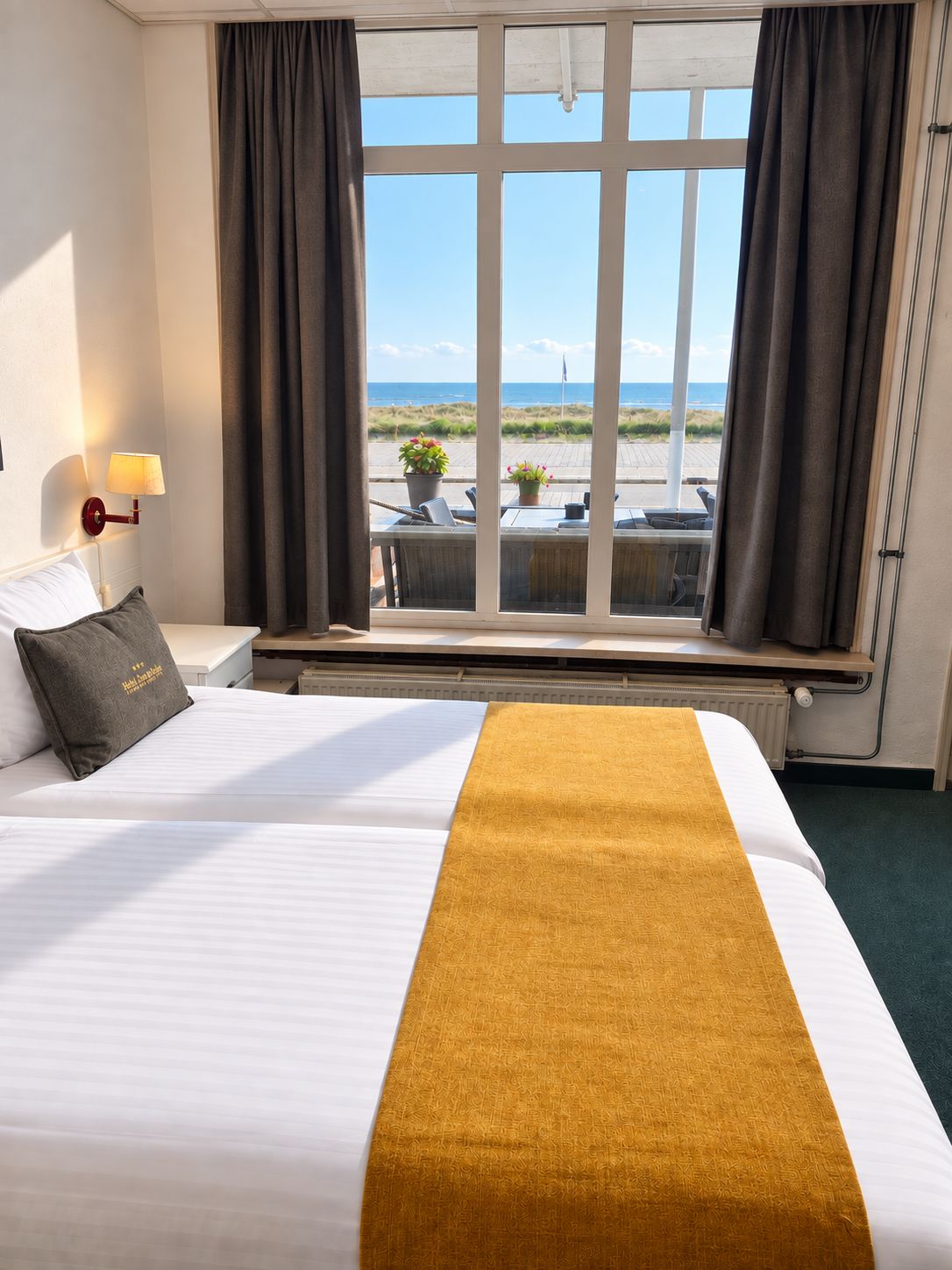 A hotel room with a bed, a nightstand, a lamp, a window with curtains, and a view of the beach.