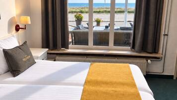A hotel room with a bed, a nightstand, a lamp, a window with curtains, and a view of the beach.