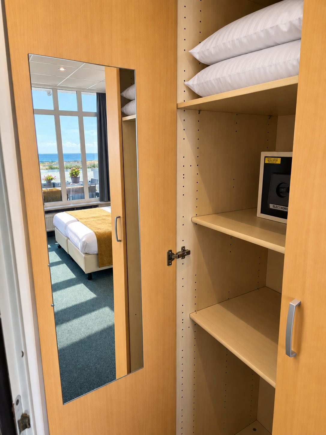 A hotel room with a bed, a mirror, and a shelf with a pillow and a small electronic device.