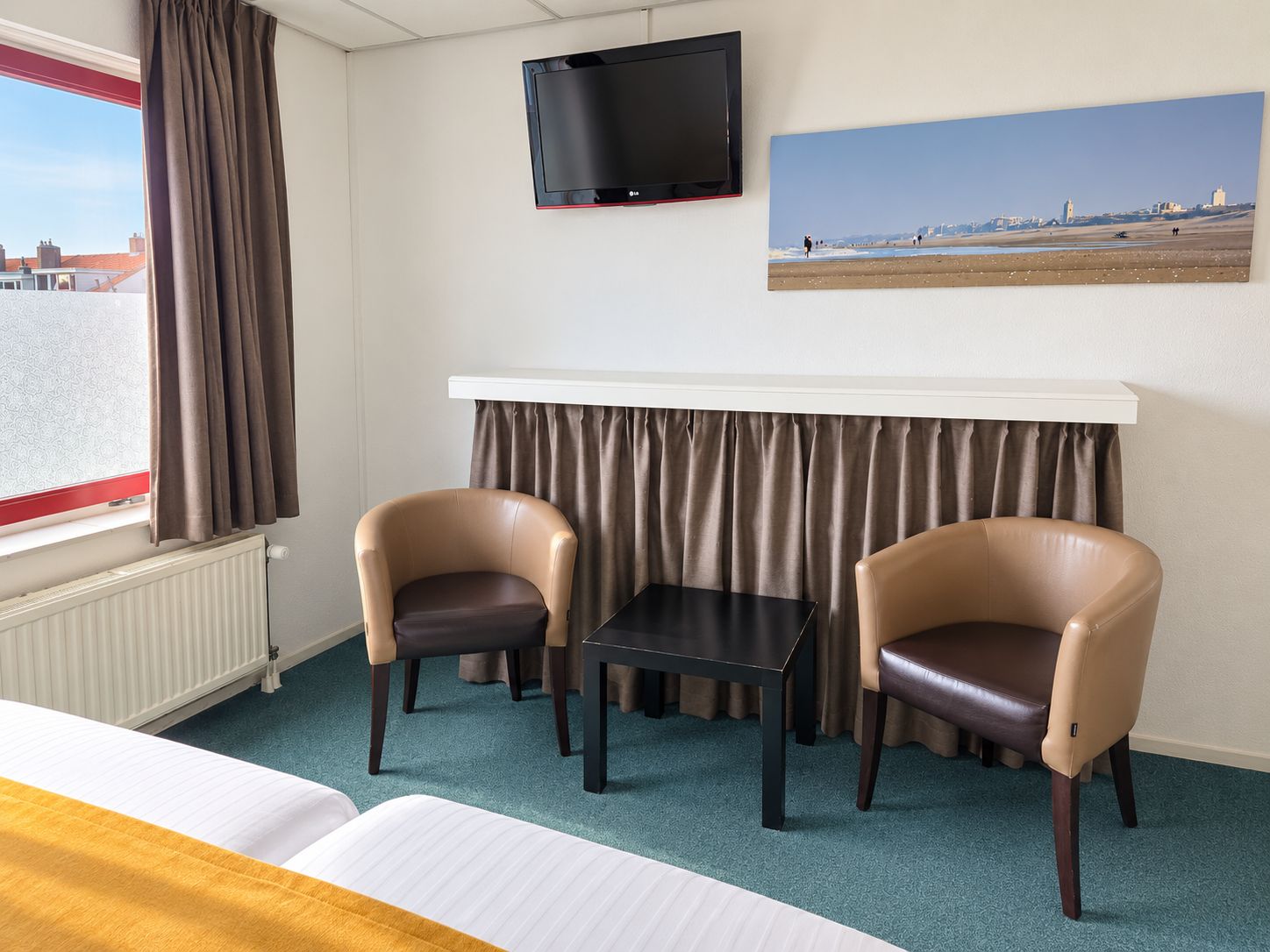 Habitación de hotel con cama, dos sillas, una mesita, televisor, un cuadro y ventana con cortinas.