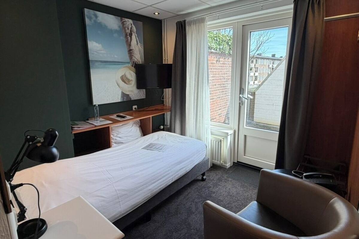 Habitación de hotel con cama, escritorio con una computadora portátil, silla, televisor y un ventanal con cortinas.