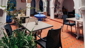Une cour intérieure avec une table et des chaises au centre, entourée d’arcades et de plantes en pot.