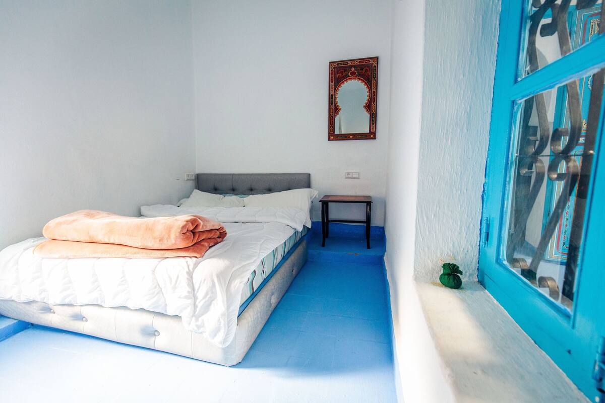 Une chambre à coucher avec un lit, une table de chevet, un miroir et une fenêtre dont les bords sont peints en bleu.