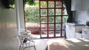 A bedroom with a bed, a small kitchenette, a window with a view of greenery, and a chair near the window. - 8 Soenie Avenue Self Catering (Akasia)
