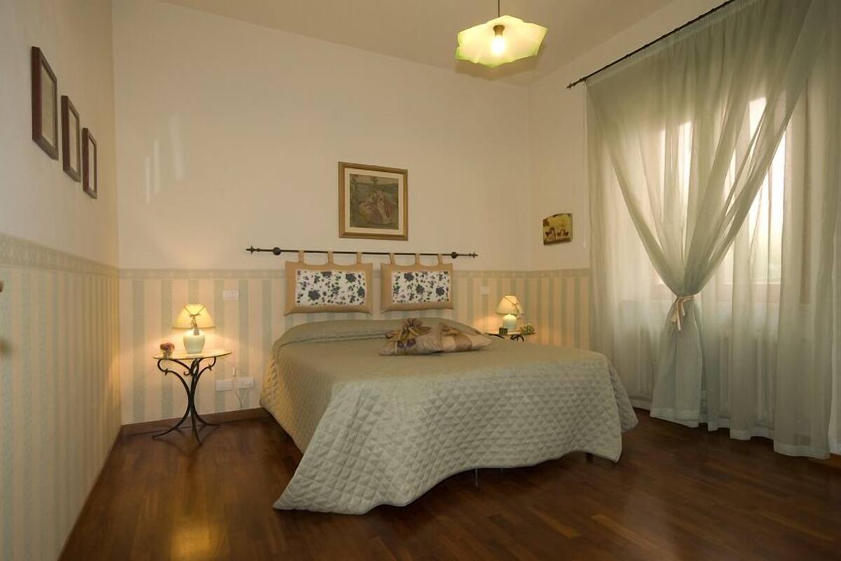 Une chambre à coucher avec un lit, deux tables de chevet avec des lampes, une fenêtre avec des rideaux légers et un tableau accroché au mur.