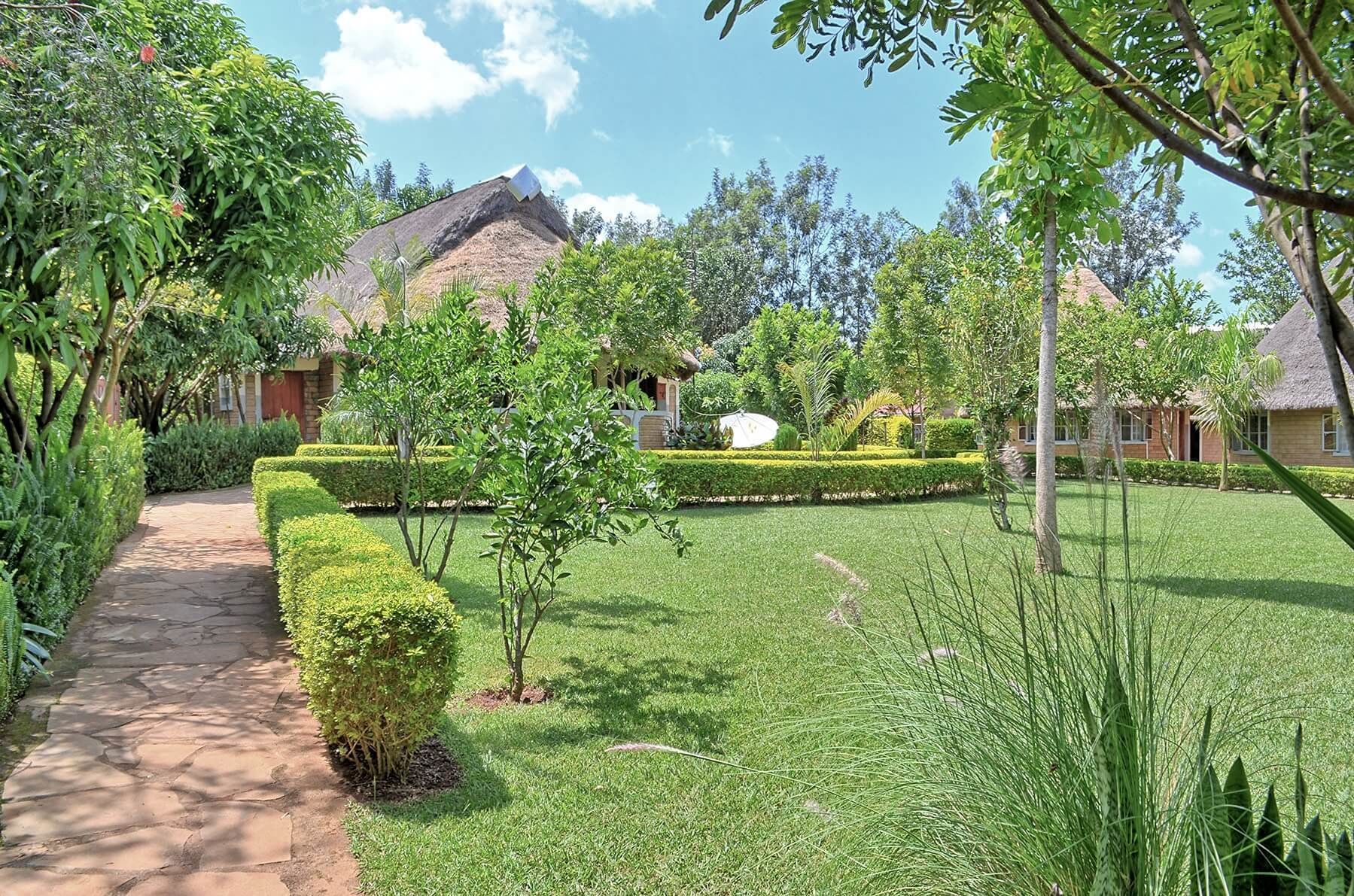 A pathway leading to a thatched-roof building surrounded by well-maintained greenery and a clear sky.