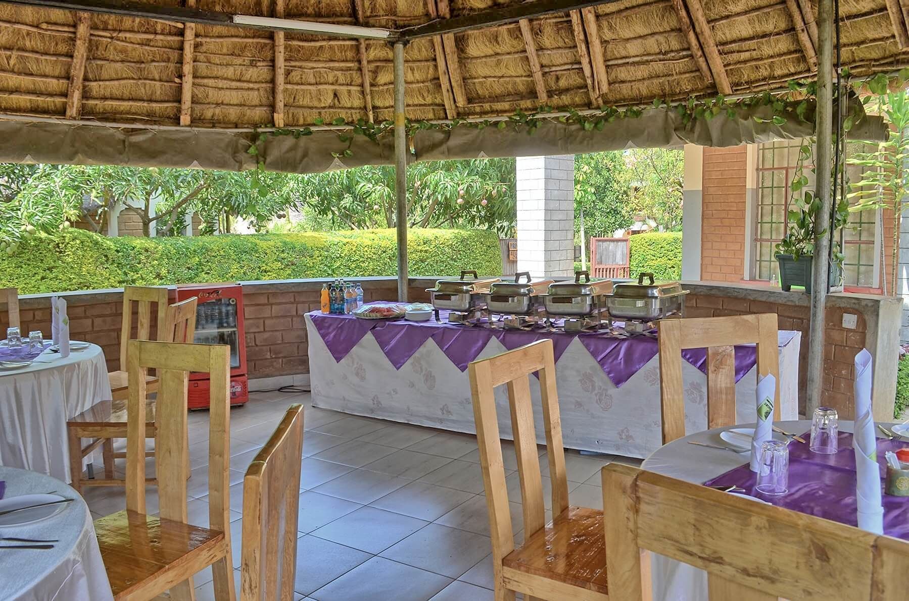 An outdoor dining area with wooden chairs and tables, a buffet setup, and a thatched roof.