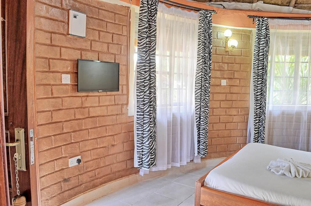 A room with a brick wall, a flat-screen TV mounted above a door, zebra-striped curtains, and a bed with white bedding.