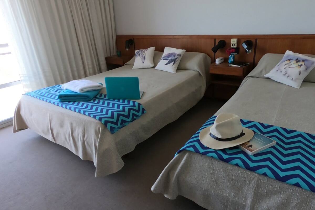 Habitación de hotel con dos camas, cada una con una colcha azul y blanca a cuadros en chevron, un sombrero blanco y un libro sobre una de las camas.