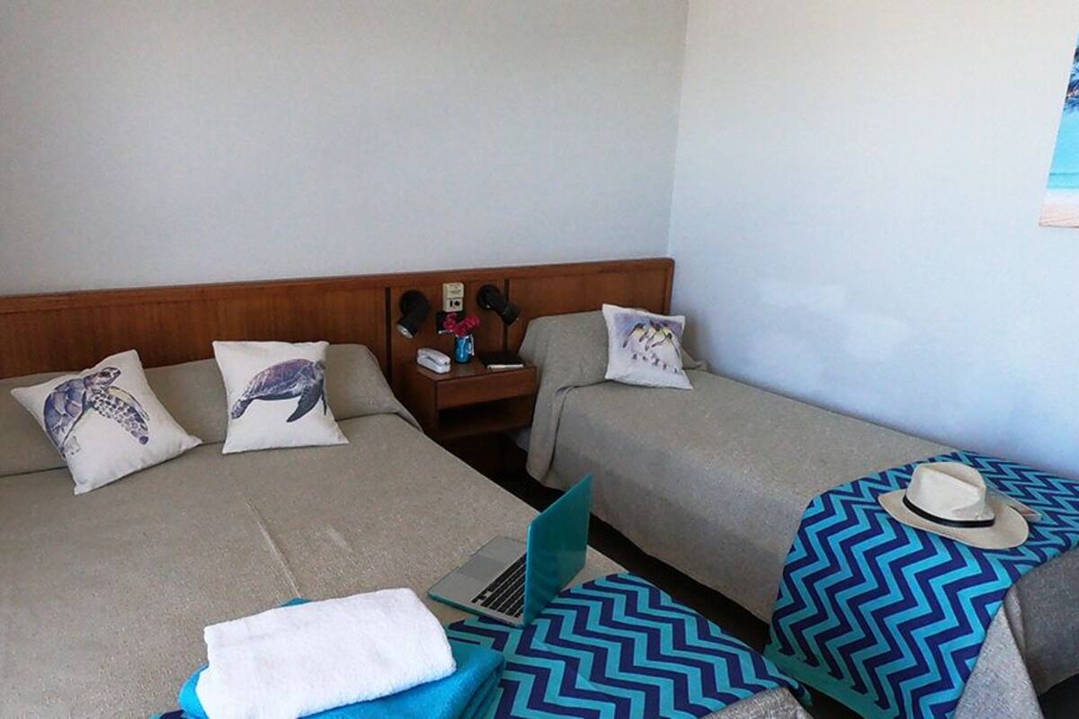 Habitación de hotel con dos camas, cada una con un cojín con diseño de tortuga. Hay un ordenador portátil en el suelo, una toalla y un sombrero sobre la cama de la derecha.