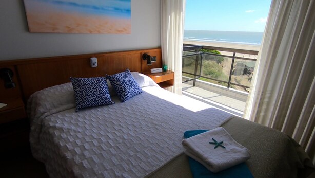 A hotel room with a large bed, two patterned pillows, a nightstand, a bedside table with a phone, a window with a view of the beach, and a towel with a star design. - Hotel Solmar (San Clemente)