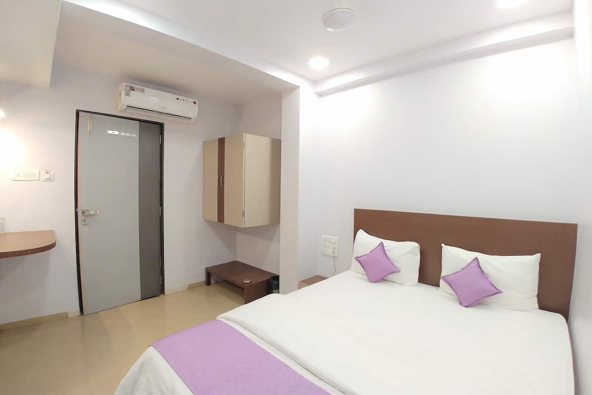 A hotel room with a bed, a wardrobe, a bedside table, an air conditioning unit, and a wall-mounted light fixture.