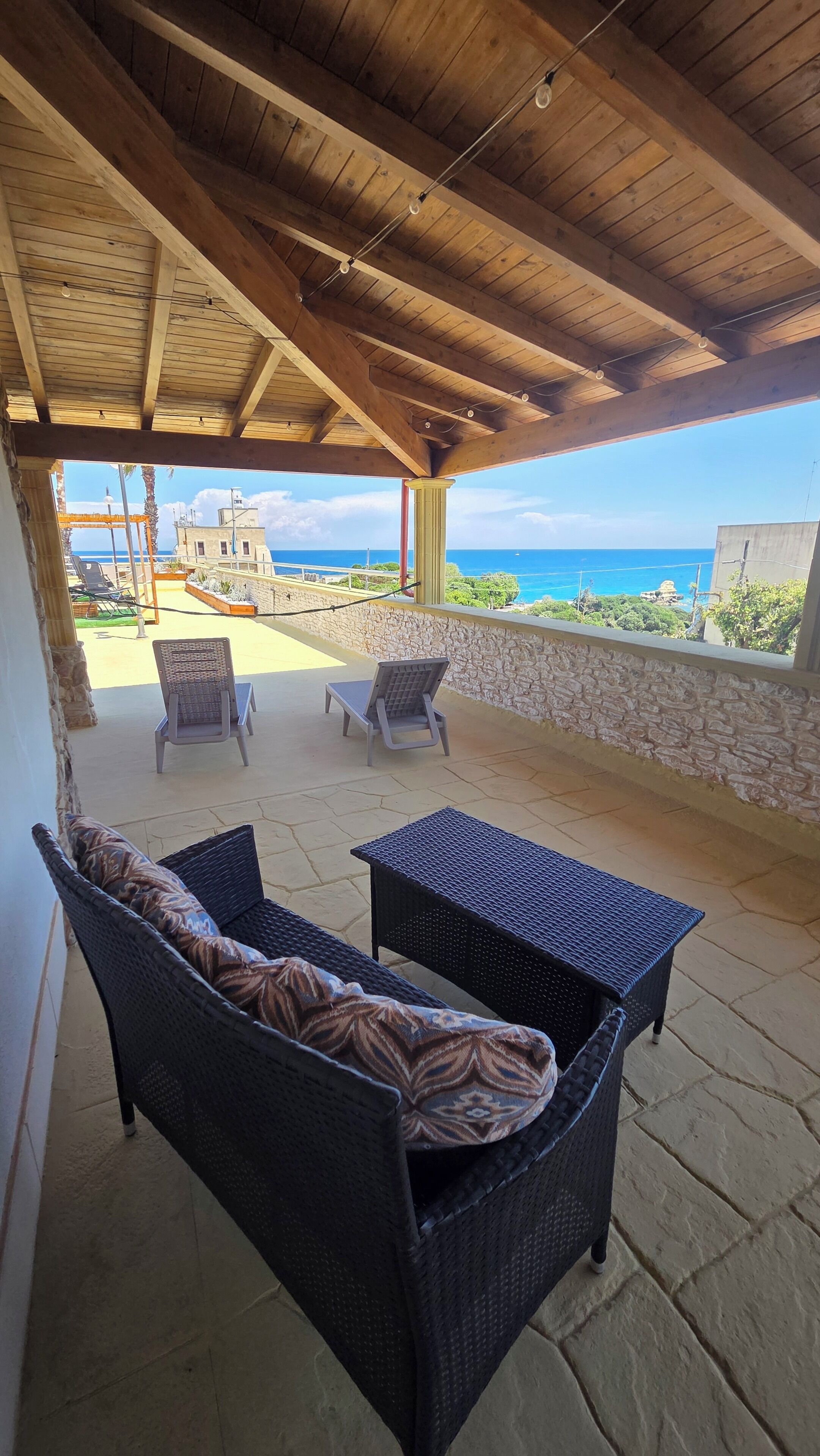 A covered outdoor seating area with wicker furniture, a stone wall, and a view of the ocean.