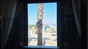 Una ventana con vista a una palmera, un edificio pequeño y coches aparcados.