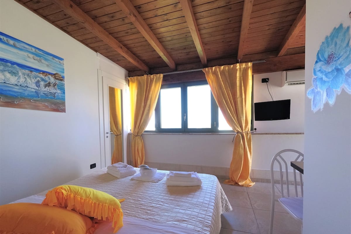 A bedroom with a wooden ceiling, a bed with white bedding and orange pillows, a chair, a television on the wall, and a window with curtains.