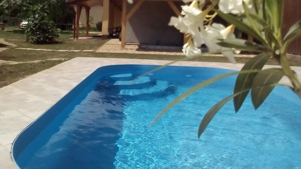 Ein Swimmingpool mit glatter, blauer Wasseroberfläche, umgeben von einem Garten mit weißen Blüten und einer hölzernen Pergola im Hintergrund.