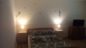 Ein Schlafzimmer mit einem Bett, zwei Nachttischen mit Lampen, einem Fernseher auf einer Holzkommode sowie Wandbildern von Schmetterlingen und Blumen.