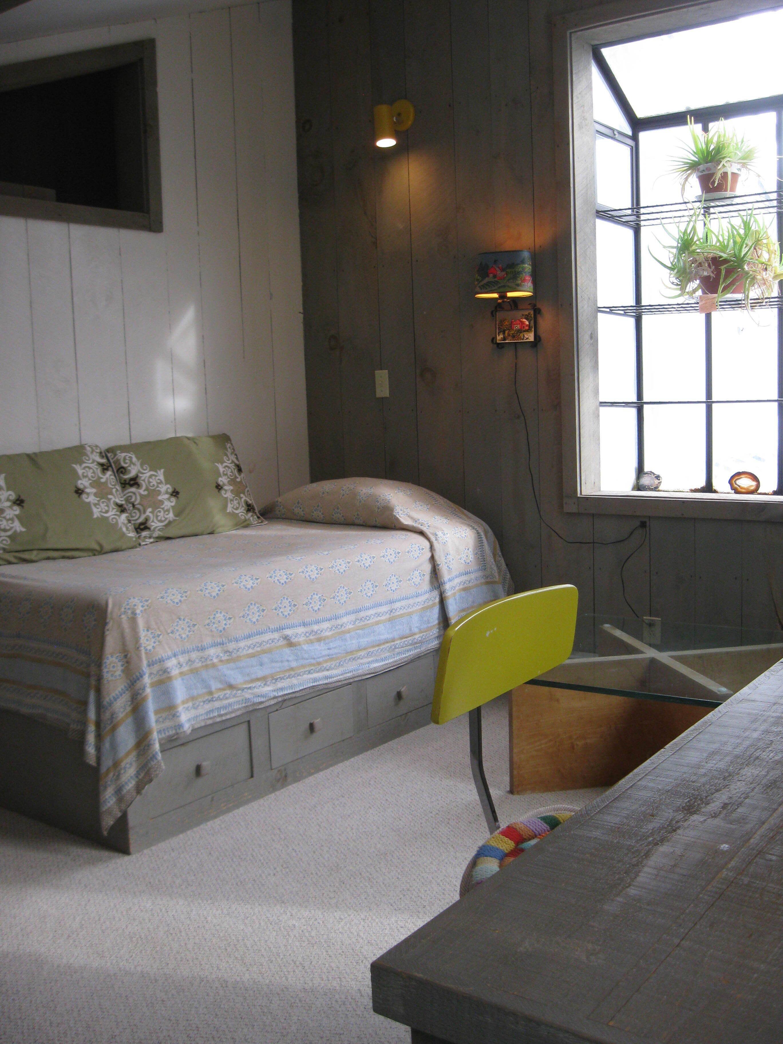 A small, cozy room with a bed, a chair, a desk, and a window with plants.