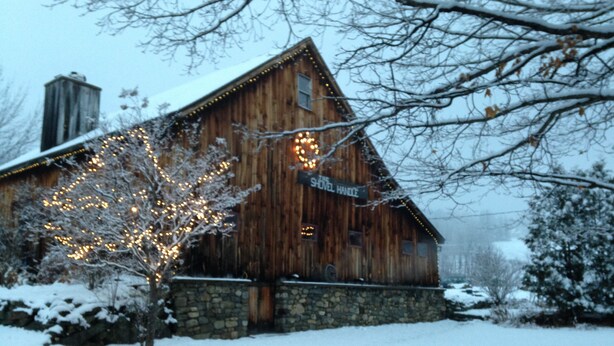 Une cabane en bois, avec des fondations en pierre, une cheminée et une enseigne sur laquelle on peut lire : « The Shovel Handle ».