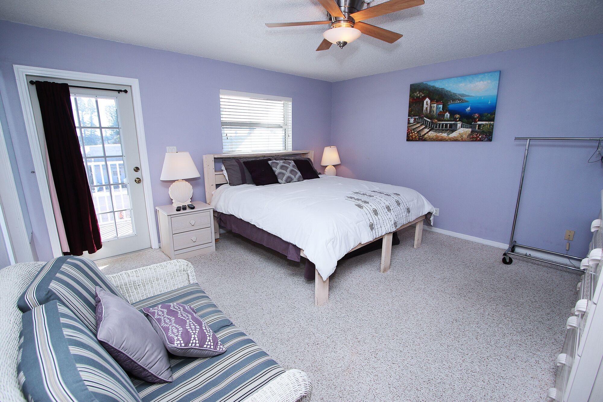 A bedroom with a bed, two bedside tables, a nightstand, a window with blinds, a ceiling fan, a painting on the wall, a standing lamp, a sofa with cushions, and a floor lamp.