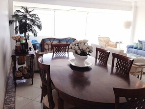A round wooden dining table with a white floral centerpiece, surrounded by wooden chairs. A potted plant and a small shelf with bottles and snacks are visible to the left. - Excellent beachfront apartment, two blocks from the beach. (Torres)