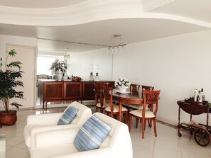 A spacious living room with a white sofa, dining table, and chairs. A kitchen area is visible in the background. - Excellent beachfront apartment, two blocks from the beach. (Torres)