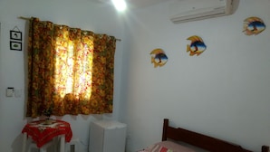 Ein Schlafzimmer mit einem Bett, einem kleinen Tisch mit roter Tischdecke, einer weißen Klimaanlage und einem Fenster mit floralen Vorhängen.
