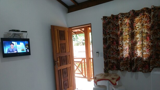 Ein kleines Zimmer mit Bett, Fernseher, der eine Nachrichtensendung zeigt, einer Holztür und einem Fenster mit Blick nach draußen.