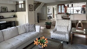 En rummelig stue med sofa, et mønstret lænestol og et sofabord med en vase blomster. En trappe fører op til en etage.