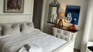 Ein ordentlich bezogenes Bett mit weißer Decke und Kissen, eine Nachttischlampe, ein dekorativer Spiegel und ein Gemälde mit einer Küstenlandschaft.