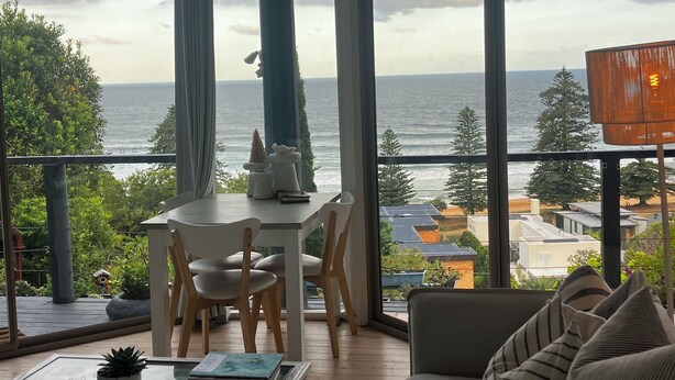 Ein modernes Wohnzimmer mit Glastisch, Sofa und Essbereich mit Stühlen. Ein großes Fenster bietet Blick auf das Meer und die umliegende Natur.