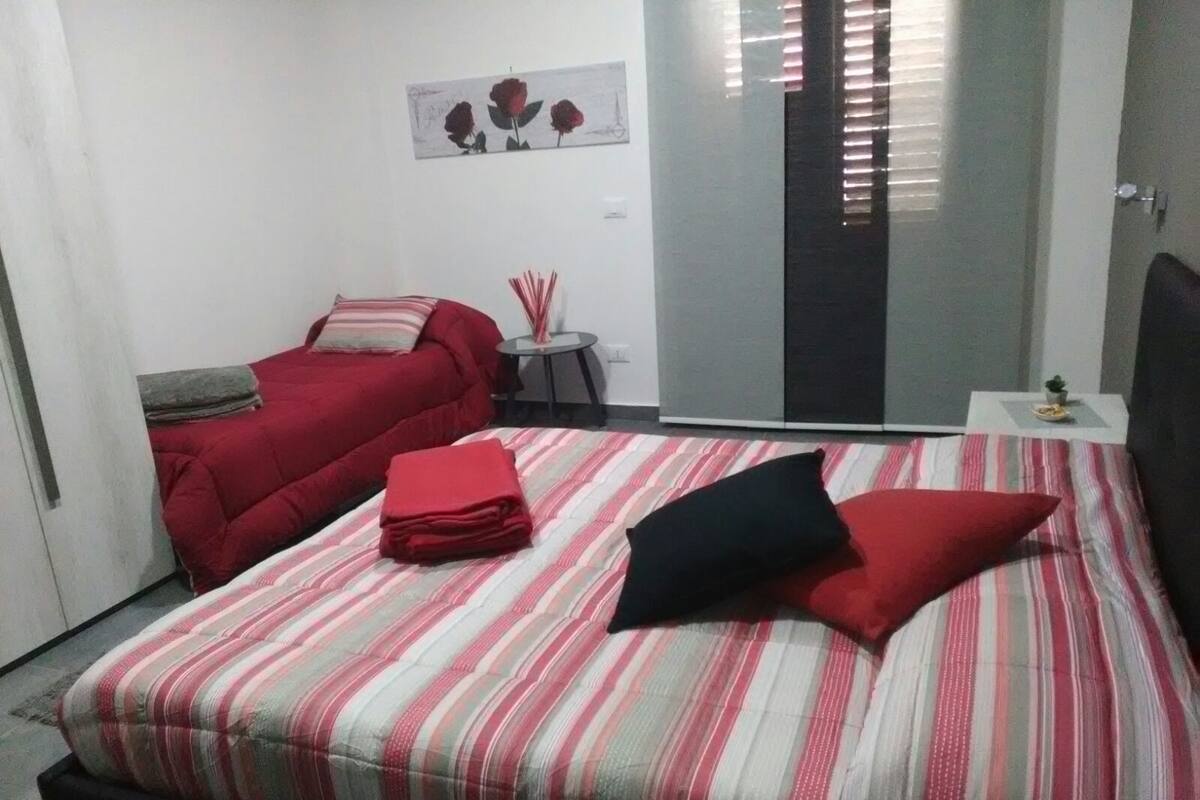 A bedroom with a bed, a red sofa, a small table with a vase, and a sliding door.