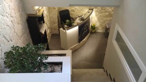 Un mostrador de recepción con un fondo de pared de piedra, una escalera que sube a un nivel superior y una planta en una maceta en el nivel inferior.
