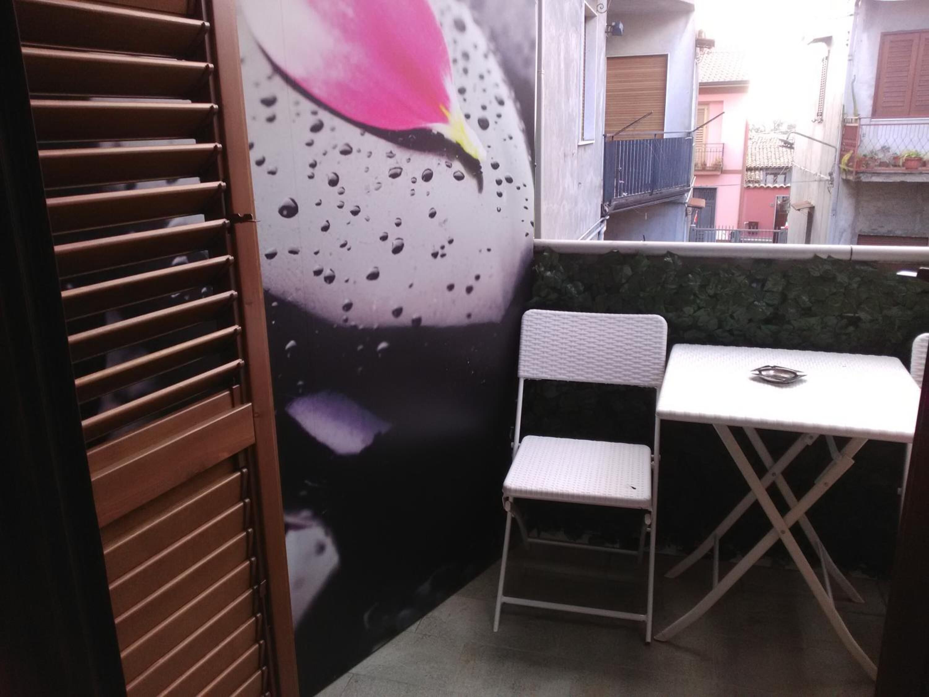 A small outdoor seating area with a white table and chair, a decorative wall with a flower, and a metal gate.
