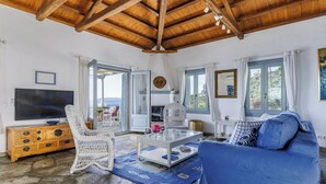 A living room with a stone floor, wooden ceiling beams, a blue sofa, wicker chairs, a glass coffee table, and a television on a wooden cabinet. - Villa Kalyves Armenopetra - Beautiful  Pool - Amazing Sea Views -Walk to Beaches (Neo Klima)